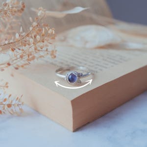 May include: A silver ring featuring a purple gemstone set in a delicate, organic design. The ring is resting on a book with a light brown cover.
