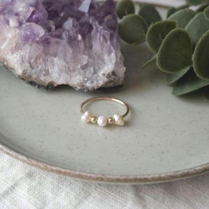 May include: A delicate gold ring featuring three white pearls and two gold beads. The ring is displayed on a light green plate, with a purple amethyst crystal and green leaves in the background. A simple and elegant piece of jewelry.