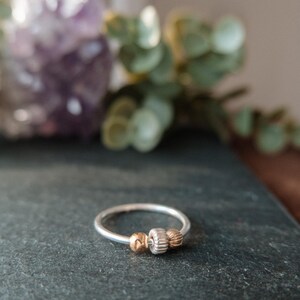 May include: A silver ring with three decorative beads. The center bead is ridged, flanked by two gold-colored beads. The ring is set against a dark gray surface, with a blurred background of purple and green.
