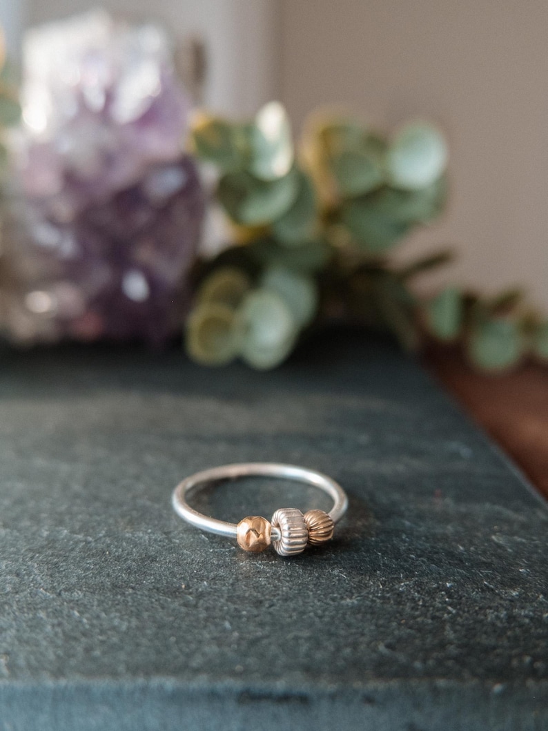 May include: A silver ring with three decorative beads. The center bead is ridged, flanked by two gold-colored beads. The ring is set against a dark gray surface, with a blurred background of purple and green.