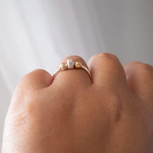 May include: A silver ring with three decorative elements: two gold-colored spheres and a textured, cylindrical silver piece. The ring is worn on a finger, showcasing the contrast between the silver band and the gold accents. The ring's design is simple yet elegant.