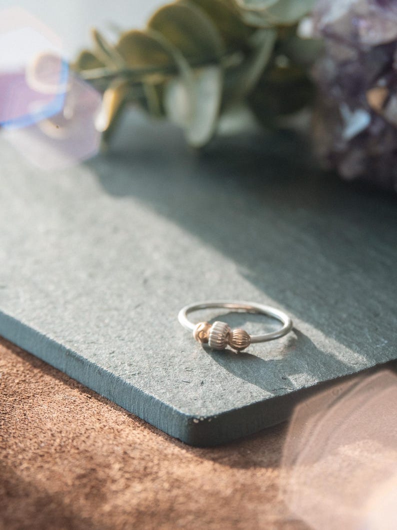 May include: A silver ring with two gold-colored beads. The ring is on a gray surface, with green leaves and purple crystals in the background. The ring is a delicate piece of jewelry.