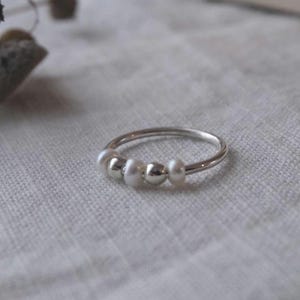 May include: A silver ring with three white pearls and two silver beads. The ring is set against a neutral background, highlighting the delicate design. This piece of jewelry is a simple yet elegant accessory.