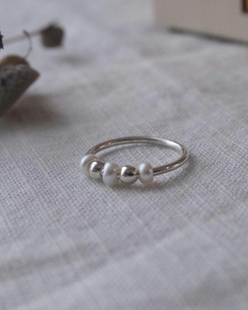 May include: A silver ring with three white pearls and two silver beads. The ring is set against a neutral background, highlighting the delicate design. This piece of jewelry is a simple yet elegant accessory.