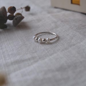 May include: A delicate silver ring featuring three small, lustrous white pearls. The ring is set against a soft, neutral background, with a hint of dried floral accents in the blurred background. A simple and elegant piece of jewelry.