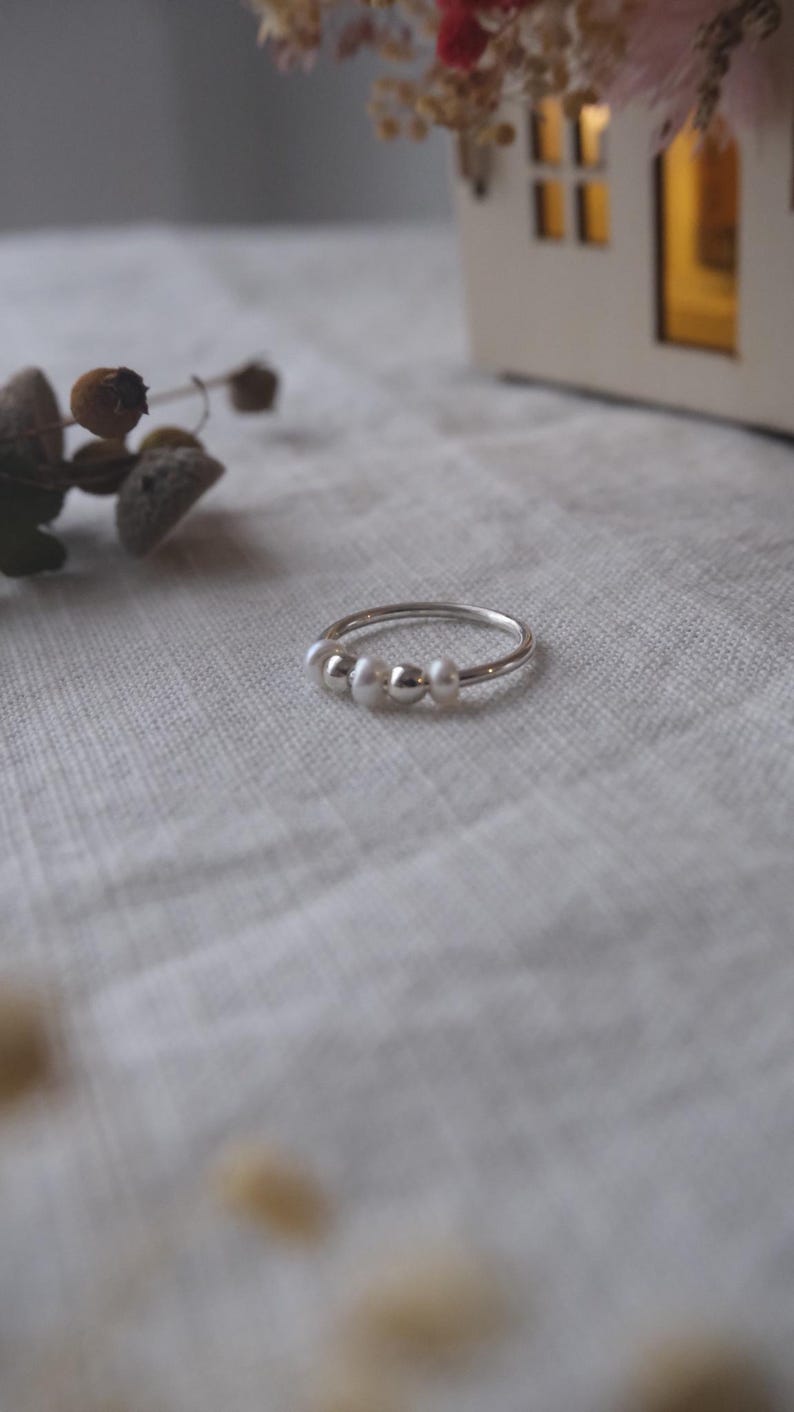 May include: A delicate silver ring featuring three small, lustrous white pearls. The ring is set against a soft, neutral background, with a hint of dried floral accents in the blurred background. A simple and elegant piece of jewelry.