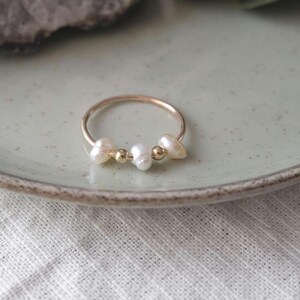 May include: A delicate gold ring featuring three white freshwater pearls and two small gold beads. The ring is set on a light green ceramic plate, with a white fabric in the background.