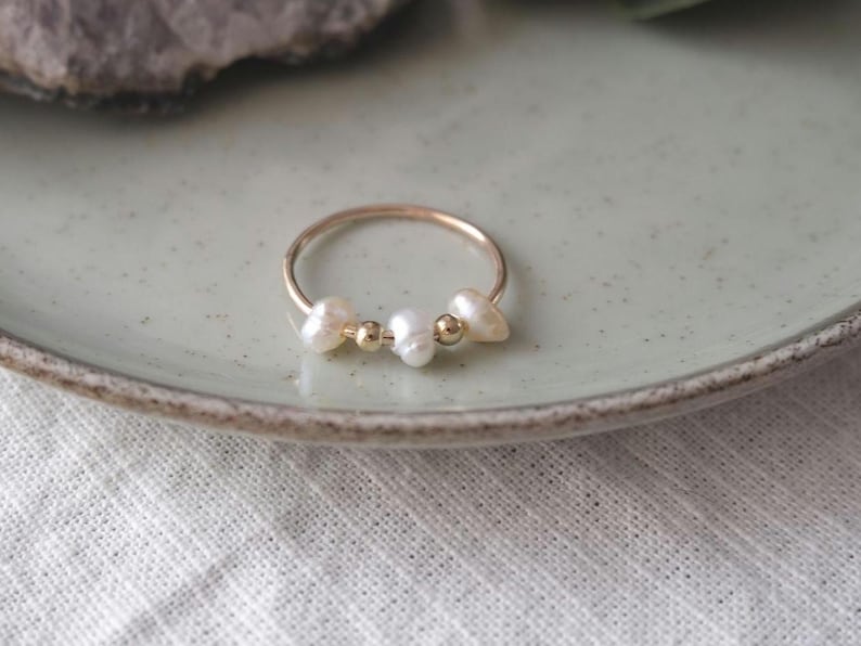 May include: A delicate gold ring featuring three white freshwater pearls and two small gold beads. The ring is set on a light green ceramic plate, with a white fabric in the background.