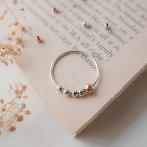 Puede incluir: Un anillo de plata con una cuenta dorada y cinco cuentas de plata más pequeñas. El anillo está sobre un libro con texto.