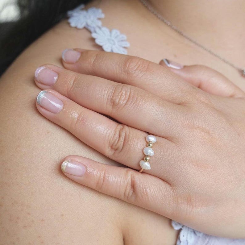 May include: A delicate gold ring featuring three freshwater pearls and small gold beads. The ring is worn on a hand with manicured nails, showcasing a minimalist and elegant jewelry design.