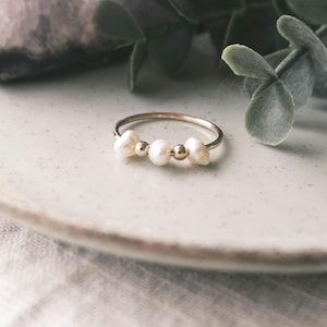 May include: A delicate silver ring featuring three freshwater pearls and two small gold beads. The ring is displayed on a light-colored plate, with greenery in the background. A simple and elegant piece of jewelry.