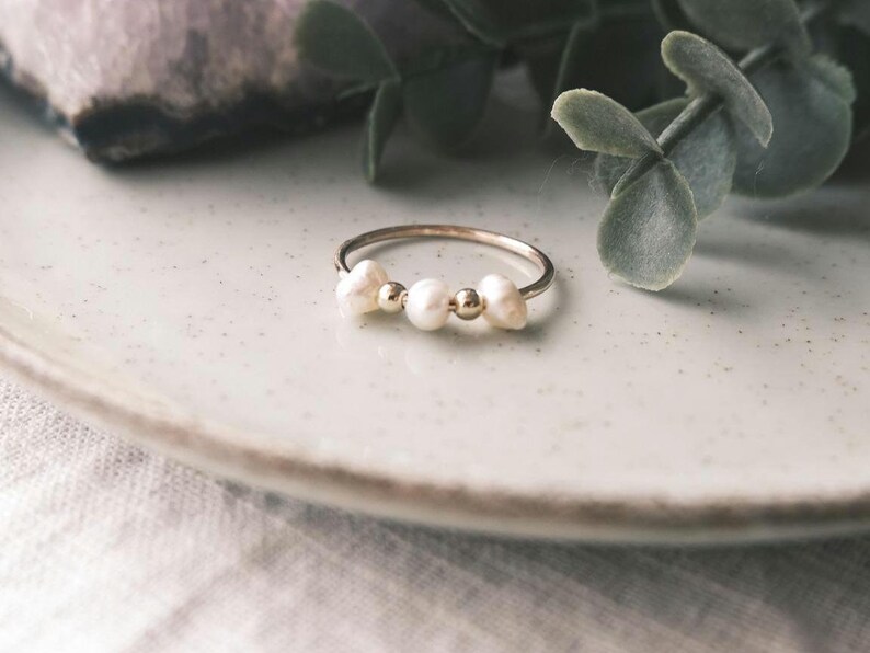 May include: A delicate silver ring featuring three freshwater pearls and two small gold beads. The ring is displayed on a light-colored plate, with greenery in the background. A simple and elegant piece of jewelry.