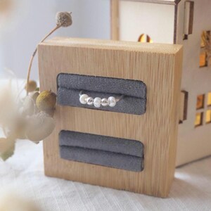 May include: A silver ring with four small, round beads displayed on a wooden jewelry holder with two gray velvet slots. A small, illuminated wooden house is in the background. The ring is the focal point.