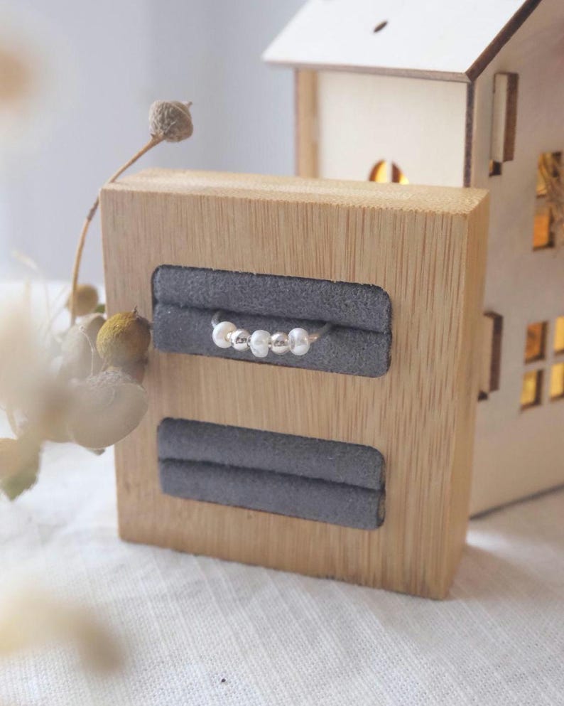 May include: A silver ring with four small, round beads displayed on a wooden jewelry holder with two gray velvet slots. A small, illuminated wooden house is in the background. The ring is the focal point.