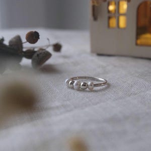 Könnte beinhalten: Ein zarter Silberring mit drei kleinen, runden, weißen Perlen. Der Ring ist vor einem weichen, neutralen Hintergrund platziert, mit einem verschwommenen dekorativen Element im Hintergrund. Ein schlichtes, elegantes Schmuckstück.