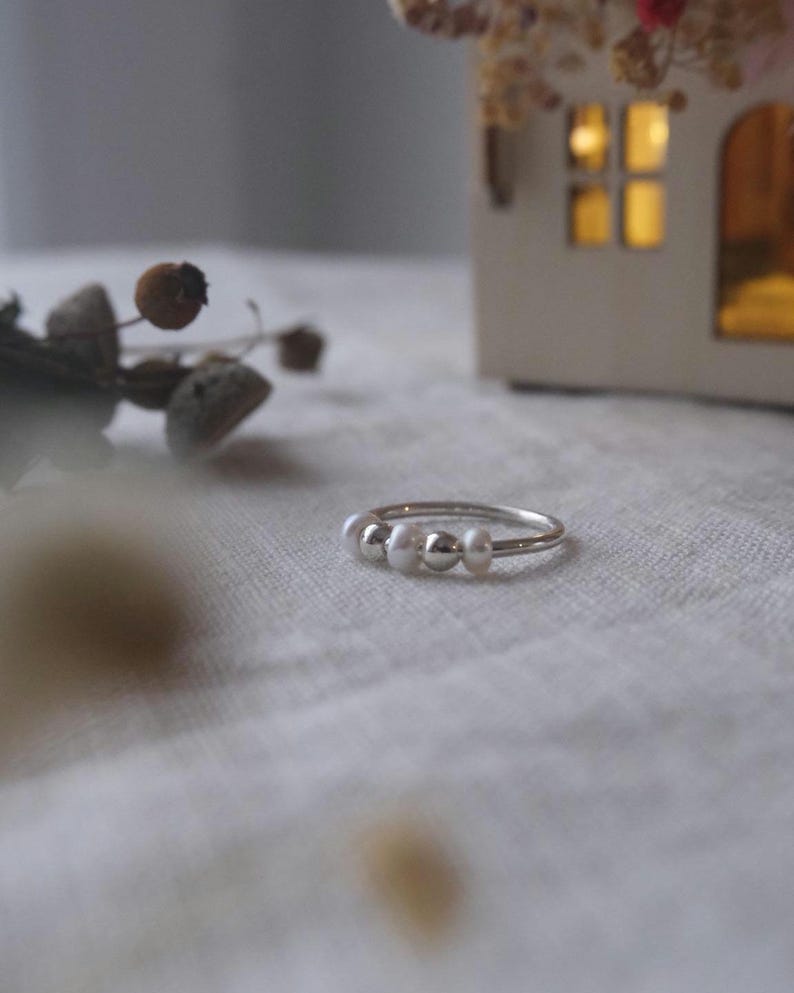 May include: A delicate silver ring featuring three small, round, white pearls. The ring is set against a soft, neutral background, with a blurred decorative element in the background. The ring is a simple, elegant piece of jewelry.