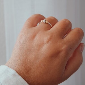 May include: A silver ring with three decorative beads, two gold and one white, is worn on a hand. The ring is simple and elegant, with a minimalist design. The background is a soft, neutral color.