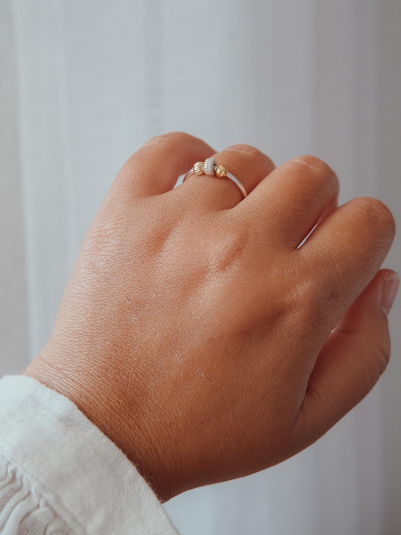 May include: A silver ring with three decorative beads, two gold and one white, is worn on a hand. The ring is simple and elegant, with a minimalist design. The background is a soft, neutral color.