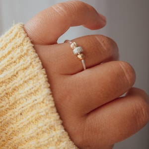 May include: A silver ring with gold and silver beads. The ring is on a finger, with a yellow sweater in the background. The ring has a simple design, with a thin band and small beads.
