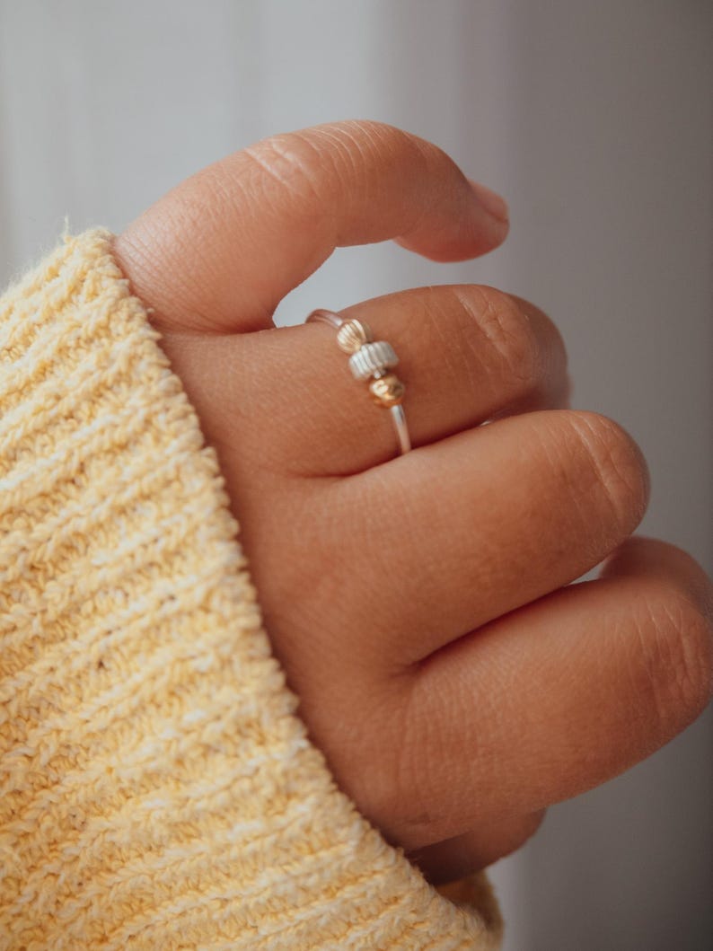 May include: A silver ring with gold and silver beads. The ring is on a finger, with a yellow sweater in the background. The ring has a simple design, with a thin band and small beads.