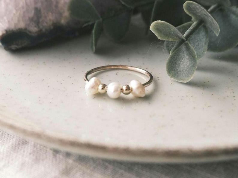 May include: A delicate silver ring featuring three white pearls and two small gold beads. The ring is set against a soft, neutral background, highlighting the jewelry's simple elegance. The pearls have an irregular shape, adding a touch of natural beauty.