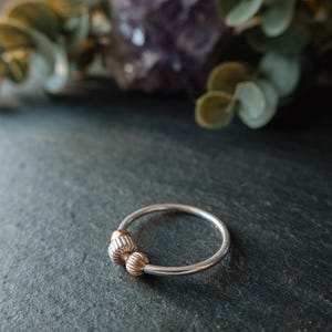May include: A silver ring with two gold-colored, textured beads. The ring is set against a dark, textured surface, with blurred greenery and a purple crystal in the background. The ring is a piece of jewelry.