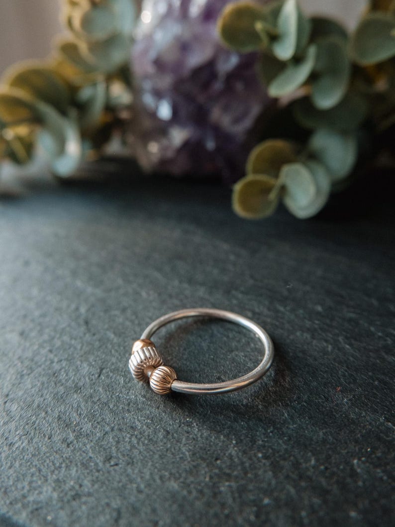 May include: A silver ring with two gold-colored, textured beads. The ring is set against a dark, textured surface, with blurred greenery and a purple crystal in the background. The ring is a piece of jewelry.