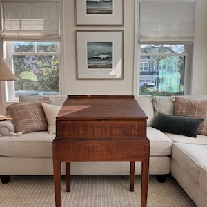 May include: A wooden writing desk with a slanted top and a small drawer. The desk is a warm brown color and stands on four slender legs. Above the desk, two framed watercolor paintings of ocean waves hang on the wall. The room has large windows and a neutral color palette.