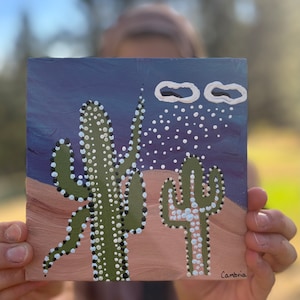 May include: A painting of two cacti with white dots on a purple background with white clouds and white dots. The painting is signed "Cambria" in the bottom right corner.