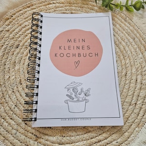 Pode incluir: Um caderno de espiral com capa branca apresentando um círculo rosa, o texto "MEIN KLEINES KOCHBUCH" com um símbolo de coração, um desenho de linha de uma panela com vegetais e o texto "OUR.BUDGET.COUPLE" impresso na borda inferior.