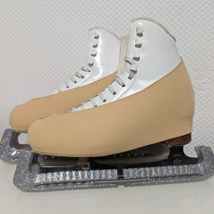May include: A pair of white figure skates with tan fabric covers over the blades. The skates have silver glitter guards on the blades.