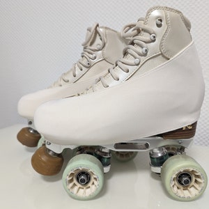 Może przedstawiać: Para białych rolek z beżowymi osłonami na buty. Rolki mają srebrną ramę i zielone kółka.