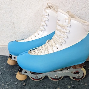 May include: White roller skates with blue covers and white laces. The skates have four wheels and the text "SNOW WHITE" is printed on the bottom of the right skate.