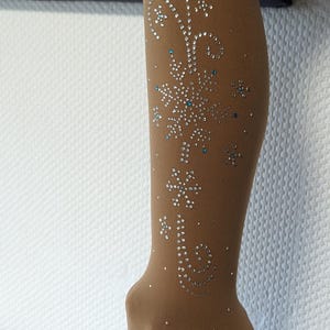 Peut inclure: Collants couleur chair avec un motif scintillant représentant un flocon de neige et un motif en spirale. Les collants sont portés avec des patins à roulettes blancs.