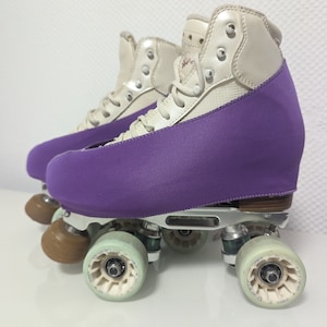 Op de afbeelding: Een paar witte rol schaatsen met paarse laarsbeschermers. De schaatsen hebben bruine wielen en een zilveren frame.