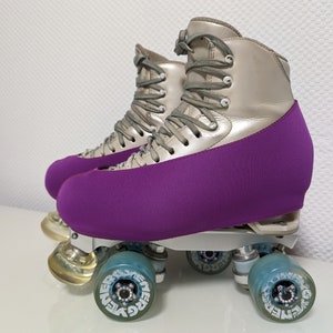 Op de afbeelding: Een paar zilveren rol schaatsen met paarse laarzenhoezen. De schaatsen hebben vier wielen met een blauw-wit ontwerp en de tekst "Energy" gedrukt op de wielen.