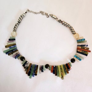 May include: A colorful necklace featuring a variety of rectangular stone beads in shades of green, blue, red, and brown. The necklace has a silver-toned chain and clasp, with decorative silver beads and a clear, round bead near the clasp.
