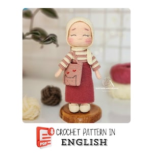 May include: Crochet pattern in English for a pink and white amigurumi doll wearing a headscarf and carrying a pink bag. The doll is standing on a wooden base.