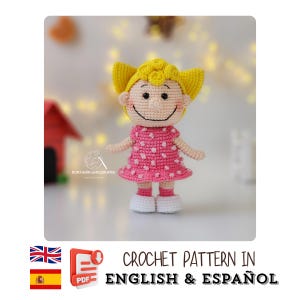 Puede incluir: Una muñeca de ganchillo con pelo amarillo y un vestido rosa con lunares blancos. La muñeca tiene una cara sonriente y zapatos blancos. La imagen incluye el texto "CROCHET PATTERN IN ENGLISH & ESPAÑOL".