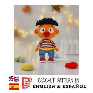 May include: A crocheted doll with an orange head, black hair, and a red nose. The doll is dressed in a striped top, blue trousers, and red shoes. The text "CROCHET PATTERN IN ENGLISH & ESPAÑOL" is displayed.