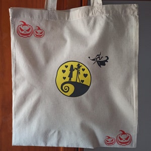 Puede incluir: Una bolsa de lona blanca con dise&ntilde;os impresos en negro y rojo. La bolsa presenta un dise&ntilde;o de Jack Skellington y Sally, dos calabazas rojas y una silueta en blanco y negro del perro de Jack Skellington, Zero.