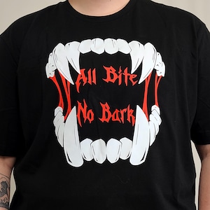 May include: A black short-sleeved t-shirt featuring a large graphic of white fangs and red saliva strands. The text "All Bite No Bark" is printed in red, stylised gothic lettering in the centre of the open mouth design on the front of the shirt.