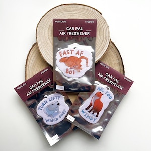 May include: Three car air fresheners with colorful designs and humorous text. The first air freshener features a cartoon cat running and the text "Fast AF Boi". The second air freshener features a cartoon dog and the text "Turn Left? Which one?". The third air freshener features a cartoon fox and the text "NC Too Wimdy".