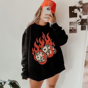 Op de afbeelding: Zwart sweatshirt met een afbeelding van twee dobbelstenen in vlammen. De dobbelstenen hebben hartsymbolen. De outfit bevat een oranje baseballcap met de tekst "Live Love Hot".