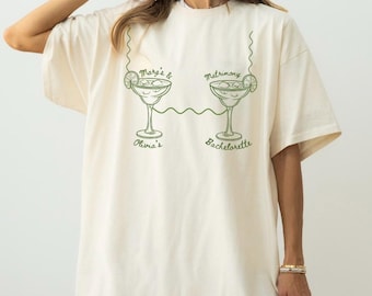 Puede incluir: Camiseta crema de gran tamaño con una ilustración verde de dos copas de margarita conectadas por una línea. El texto encima de las copas dice "Mare's & Matrimony", y debajo, "Olivia's Bachelorette".