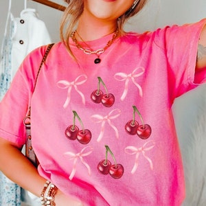 May include: A pink t-shirt with a graphic print of cherries with pink bows.