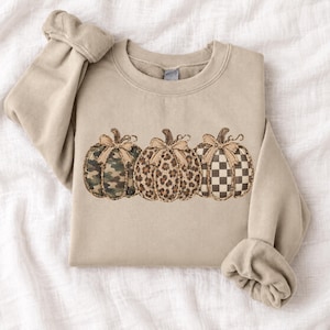 Pode incluir: Uma camisola de gola redonda bege com um gráfico de três abóboras dispostas em fila. Cada abóbora tem um padrão distinto: a primeira é camuflada, a segunda tem estampado de leopardo e a terceira tem um padrão axadrezado castanho e branco.