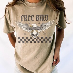 May include: A light brown t-shirt with a graphic print of an eagle with a skull in its talons. The text "FREE BIRD" is printed above the eagle. The numbers "19" and "75" are printed on either side of the eagle. A checkered pattern is printed below the eagle.