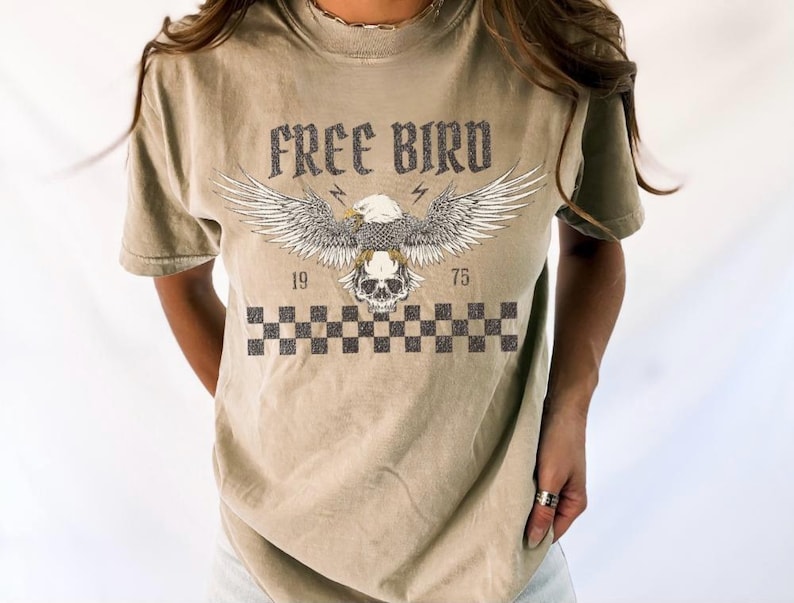 May include: A light brown t-shirt with a graphic print of an eagle with a skull in its talons. The text "FREE BIRD" is printed above the eagle. The numbers "19" and "75" are printed on either side of the eagle. A checkered pattern is printed below the eagle.