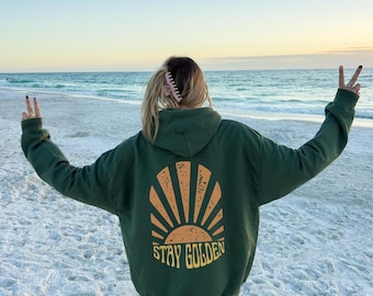 May include: A green hoodie with a yellow sun and the text "STAY GOLDEN" on the back. The person is wearing the hoodie on a beach.